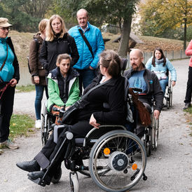 Dieses Bild zeigt Mitglieder des Projektteams beim Ausprobieren, wie sich das Naturerleben im Rollstuhl bzw. mit einem Blindenstock anfühlt. Die Begleiterin im Rollstuhl führt die Gruppe.