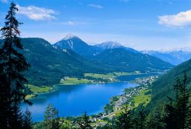 Ein Bergsee umrundet von Bergen und Wald im Sommer. Entlag des Ufers stehen Dörfer. Am Westufer des Weissensees entsteht ein 6km langer barrierefreier Weg.